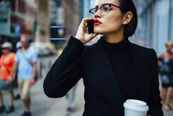 Confident female executive manager with trendy red lips talking on phone about work strolling with coffee to go, serious business woman in black elegant formal wear having mobile conversation outdoor - Foto, Imagem