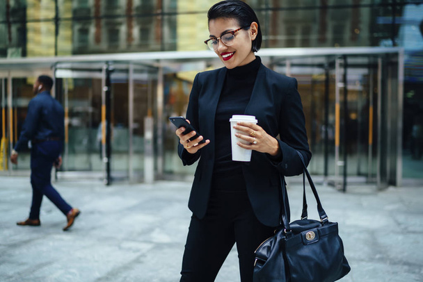 Smiling elegant businesswoman satisfied with 4G connection downloading files while standing outdoors. - 写真・画像