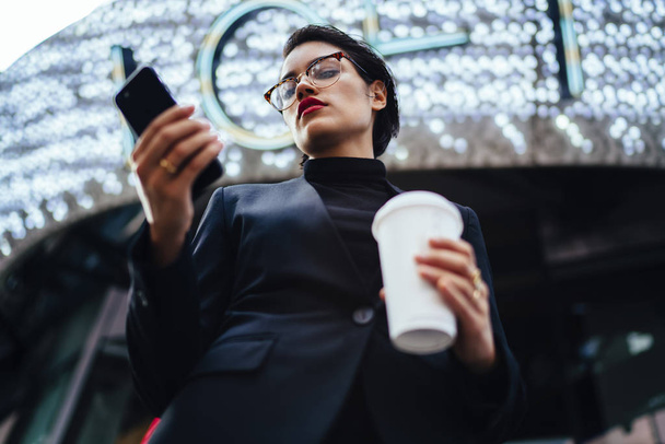 Serious female entrepreneur with red lips updating application on mobile while strolling on street with coffee to go. - 写真・画像
