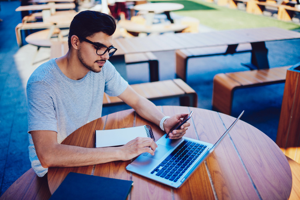 Programmatore maschio concentrato in occhiali alla moda sincronizzazione di file multimediali da cellulare con laptop in terrazza caffè
. - Foto, immagini