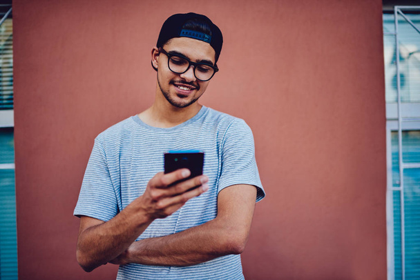Smiling hipster guy in trendy casual outfit checking email and texting feedback on smartphone standing on promotional background. - Fotografie, Obrázek
