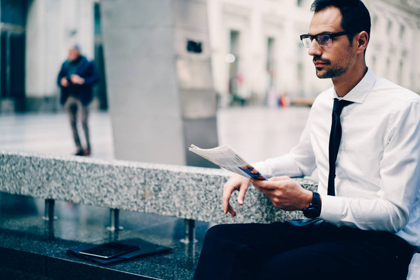 Young intelligent male economist waiting for meeting and checking some information from newspaper.Smart professional executive manager in cool spectacles looking away during sitting at urban setting - 写真・画像
