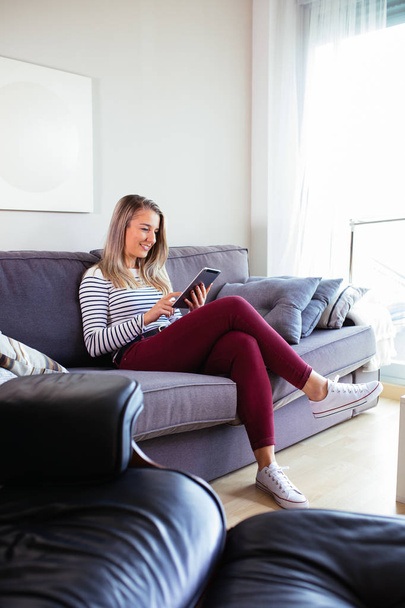 souriant jeune femme en utilisant sa tablette assis sur le canapé dans son
  - Photo, image