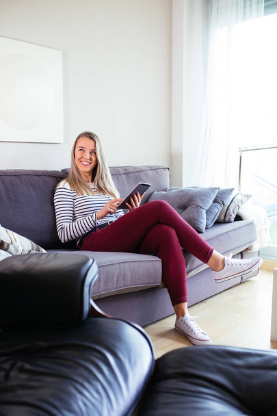 smiling young woman using her tablet sitting on the sofa in her  - Fotografie, Obrázek