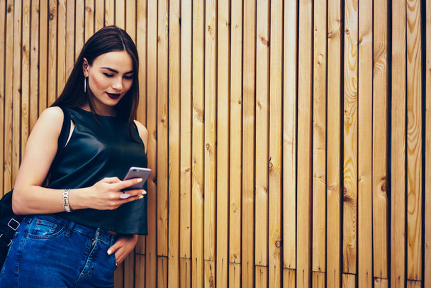 Charming young brunette dressed in trendy fashionable clothing standing against advertising area for your promotional content while sending messages to friends via smartphone connected to internet - Foto, imagen