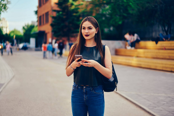 Charming brunette student strolling around city and booking online in website on modern telephone connected to 4G internet.Attractive pensive female blogger enjoying free time outdoors with gadget - Zdjęcie, obraz