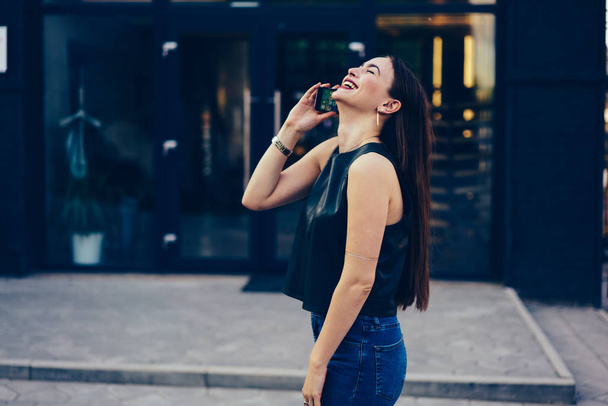 Charming emotional female blogger sincerely laughing while communicating with friends on smartphone connected to internet.Smiling brunette walking at summer street and talking on modern telephone - Фото, изображение