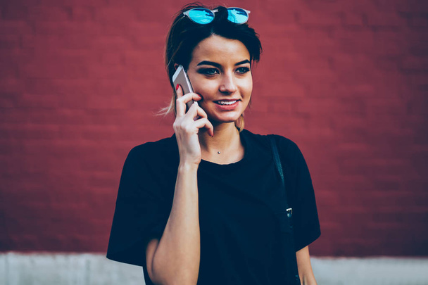 Cute happy female person talking on modern telephone during summer walk next to colorful wall with advertising area.Positive blogger enjoying recreation at urban setting near to promotional background - Photo, Image