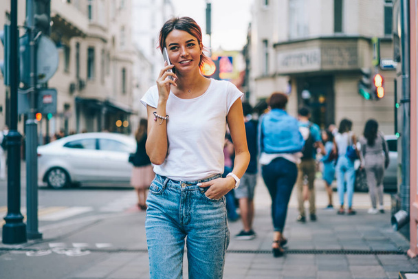 Jovem mulher elegante e alegre vestida com camiseta branca com área de mock up para mensagem de texto passeando na rua da cidade moderna e conversando com o amigo no telefone.
 - Foto, Imagem