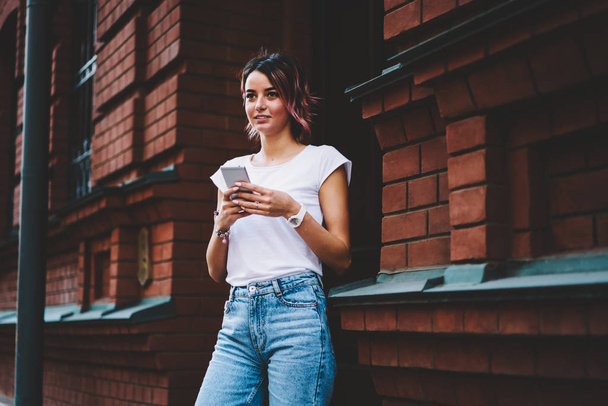 Stylish student in casual outfit checking email on modern telephone standing outdoors near university.Attractive young woman making payment online on smartphone device while looking straight - Photo, image