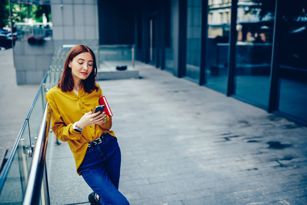 Pensive good looking woman dressed in trendy brand clothing checking email on smartphone while standing outdoors.Thoughtful cute female texting feedback in webpage via modern telephone outside - Foto, imagen