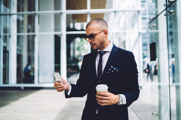 Confident male entrepreneur in trendy sunglasses reading news from networks on mobile during  coffee break, proud CEO in formal wear holding takeaway coffee texting messages on smartphone outdoor - Foto, immagini