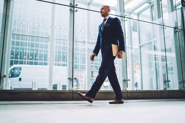 Handsome male entrepreneur in elegant suit passing glass window of modern architecture building with copy space for advertising, confident financial manager walking with documents in business center - Foto, imagen