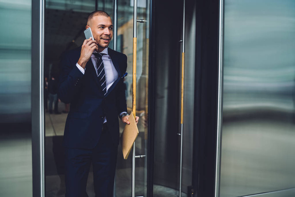 Smiling executive manager leaving building talking in phone with colleague about successful meeting, prosperous businessman in formal wear open doors sharing good news during phone conversation - 写真・画像