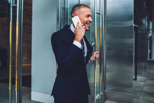 Cheerful financial manager sharing good news with colleague during phone conversation after successful meeting, positive male entrepreneur talking on mobile living office building after working day - 写真・画像