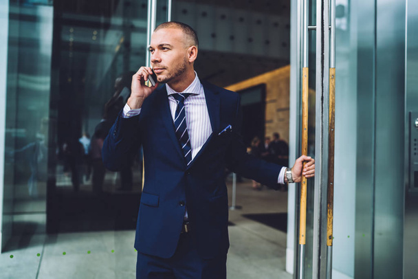 Confident male entrepreneur open door for leaving building after meeting making call to banking operator, handsome proud CEO talking on mobile with colleague standing in exit of office building - Foto, imagen