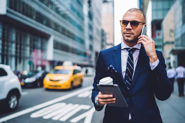Confident businessman in sunglasses talking on mobile while waiting for yellow cap on Manhattan road, serious male director holding tablet and coffee to go while calling taxi on phone getting to wor - Fotografie, Obrázek