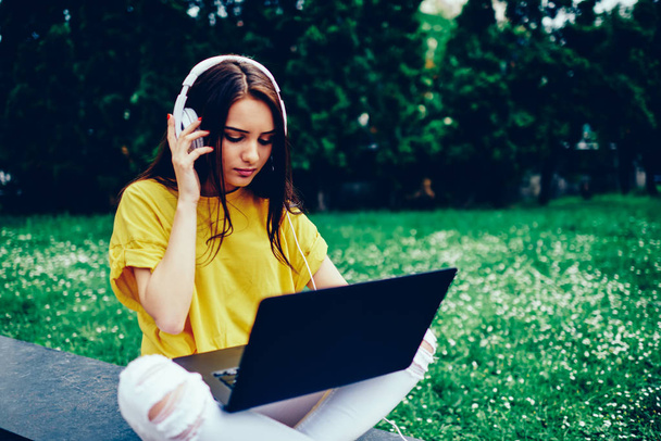 Jeune rédactrice brune talentueuse vêtue d'une tenue à la mode travaillant à distance sur un ordinateur portable connecté à Internet sans fil assis à l'extérieur.Femme réfléchissant à la recherche de musique cool sur la page Web pour écouter
 - Photo, image