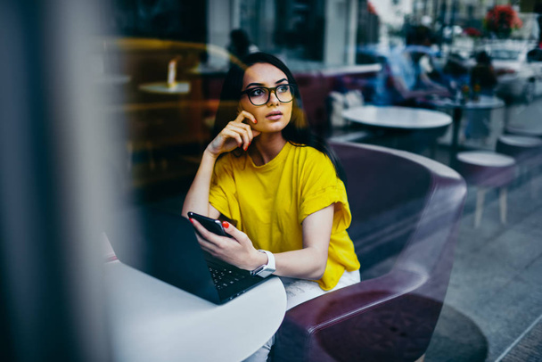 Contemplative charming graphic designer in trendy eyewear looking away and thinking about creative ideas for implementing in work.Gorgeous brunette student pondering while sitting in coffee shop - Фото, изображение