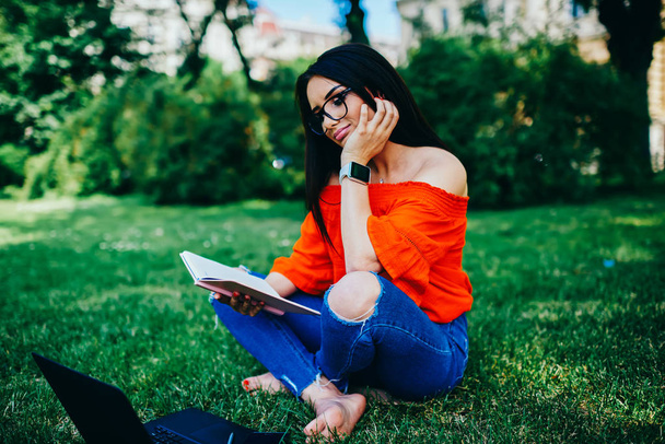 Στοχαστικός θετική hipster κορίτσι ντυμένοι με μοντέρνο ντύσιμο, κάθεται στο καταπράσινο πάρκο και Έλεγχος πληροφοριών από το Σημειωματάριο (Notepad). Ταλαντούχος φοιτήτριας προετοιμασία μαθημάτων έργου στο αστικό περιβάλλον - Φωτογραφία, εικόνα