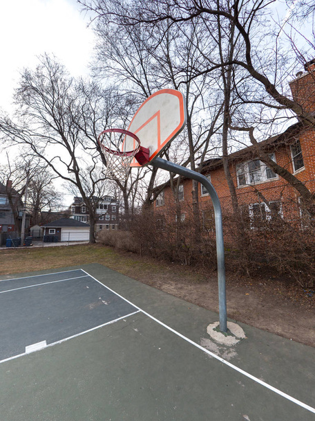 Fotografia de uma quadra de basquete urbana ao ar livre e backboard branco e laranja, borda e rede montada em um poste de metal com corte pintada de azul e verde e edifícios e árvores além
. - Foto, Imagem