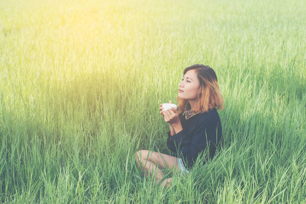 流行に敏感な若い女性がホット コーヒーを持って緑の草の上に座って、 - 写真・画像