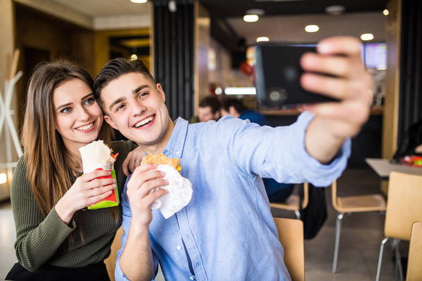 Happy smiling woman and a handsome man are enjoying their delicious and tasty burgers and doner taking selfie phone - Foto, afbeelding