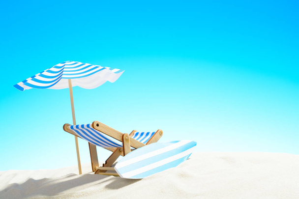 Cadeira de convés sob um guarda-chuva e uma prancha de surf na praia de areia
 - Foto, Imagem