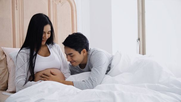 Young husband touching belly of smiling pregnant wife on bed - Fotoğraf, Görsel