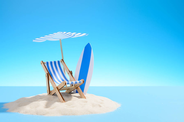 El concepto de unas vacaciones tropicales. Una chaise longue bajo un paraguas y una tabla de surf en la isla de arena. Cielo con espacio de copia
 - Foto, imagen