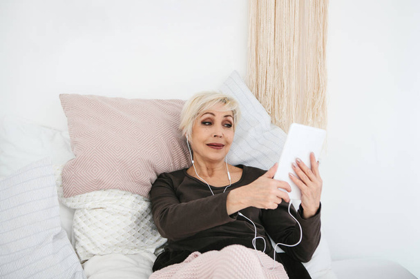 Egy idősebb pozitív nő egy tabletta segítségével videókat nézni, zenét hallgatni, és beszélgessen barátaival a szociális hálózatok. - Fotó, kép