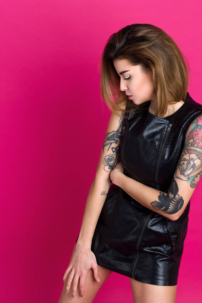 portrait of young fashion woman with tattoo on hands and leather dress on pink background - Фото, изображение
