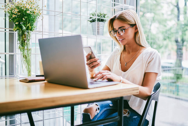 Cheerful successful female in eyeglasses received good news on smartphone while working at computer.Positive copywriter in spectacles sitting at table with laptop device and reading post on website - Φωτογραφία, εικόνα