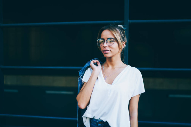 Attractive blond hipster girl in stylish optical eyeglasses looking straight while standing against promotional background.Charming young woman with trendy denim jacket on shoulder - Valokuva, kuva
