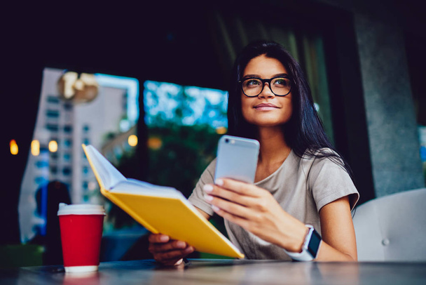 Charming young businesswoman in eyeglasses checking daily schedule in dairy and notification on smartphone on break, attractive student reading favorite book in cafe while getting message from friend - Foto, immagini
