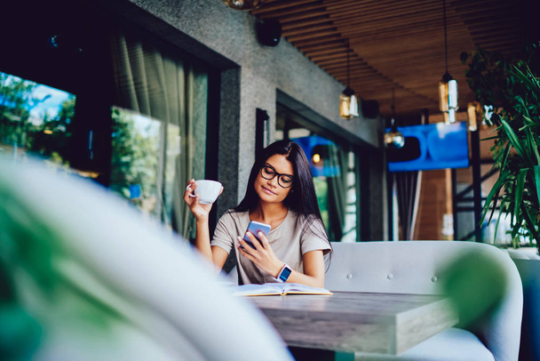 Thoughtful business woman in trendy eyewear reading financial news fro networks during morning tea time, pensive hipster girl resting on break in coffee shop viewing photo of friends on smartphone - 写真・画像