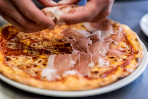 Gros plan de la main d'un chef, préparation d'une pizza base de tomate proshuto rukola et fromage râpé
 - Photo, image