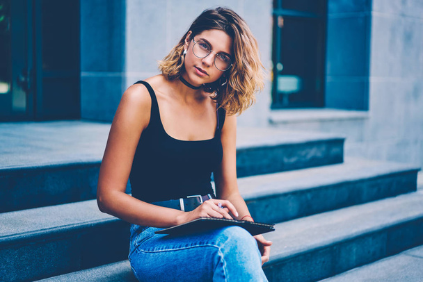 Portrait of concentrated female sitting outdoors while planning project and noting creative ideas, talented hipster girl in eyewear spending time drawing sketch in notepad  looking at camera - Foto, Imagen