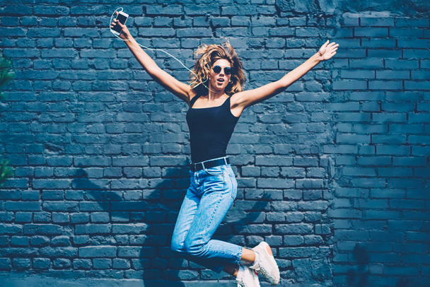 Happy young woman having fun outdoors jumping high on promotional background listening music on smartphone, carefree girl enjoying new song from playlist on mobile excited with sound making leap - Fotó, kép