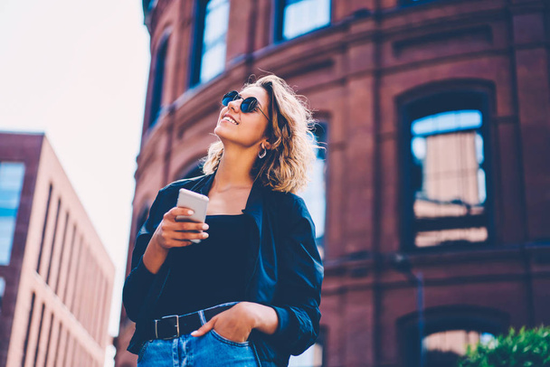 Positive hipster girl in sunglasses enjoying spring day using mobile for chatting in social networks, smiling young woman dressed in stylish outfit feeling excited with spending time outdoors - Foto, imagen