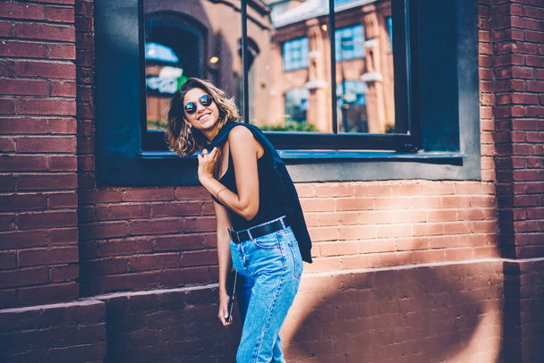 Happy young female in sunglasses having fun while spending time outdoors feeling carefree during leisure, cheerful hipster girl taking off jacket enjoying warm street weather while strolling - Fotoğraf, Görsel