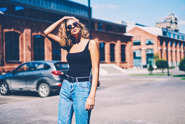 Gorgeous woman in sunglasses touching hair posing outdoors near copy space area for advertising, beautiful hipster girl dressed in cool trendy apparel strolling on city street resting on spring day - Φωτογραφία, εικόνα