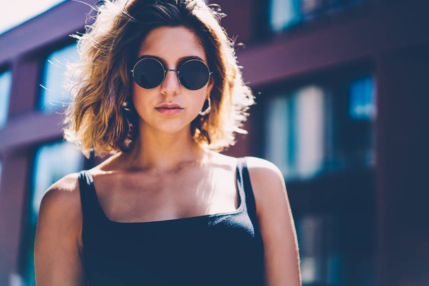 Half-length portrait of gorgeous young woman in sunglasses standing outdoors near publicity area, serious hipster girl in stylish outfit looking at camera posing on urban settings background - Foto, imagen