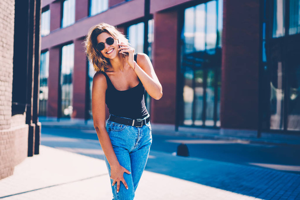 Portrait of cheerful young female in casual apparel having fun outdoors talking on mobile with friend, happy hipster girl in sunglasses excited with making calls using cheap tariffs in roaming - Photo, Image