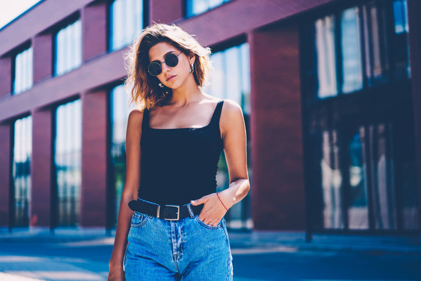 Portrait of serious hipster girl in stylish apparel standing on city street near copy space area for advertising,charming young woman in sunglasses resting outdoors during spring day looking at camera - Foto, Bild