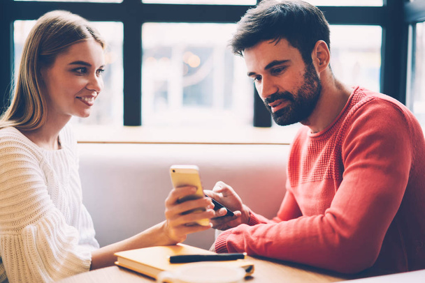 Happy young hipster girl showing her boyfriend web store site making shopping together via application on smartphone, couple in love spending time together searching restaurant for dating online - Foto, Imagen