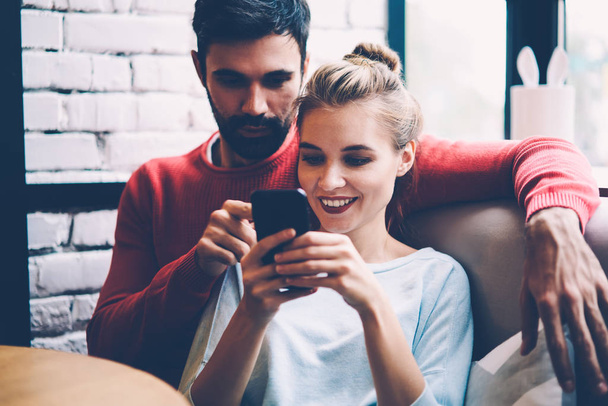 Happy romantic couple in love chatting with common friends while waiting for meeting in cozy coffee shop, smiling hipster girl viewing photos online with her boyfriend choosing best one for share - Photo, Image
