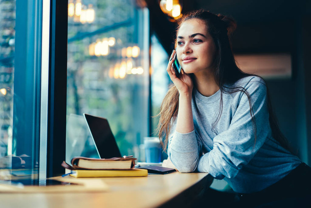 Charming positive teenager looking away while having mobile conversation with friends sitting indoors.Beautiful female blogger enjoying leisure time in coffee shop during talking on telephone - Photo, Image