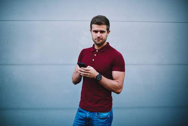 Portrait of handsome young man dressed in casual outfit looking at camera while holding modern telephone in hand and chatting online with friends standing on promotional background - 写真・画像