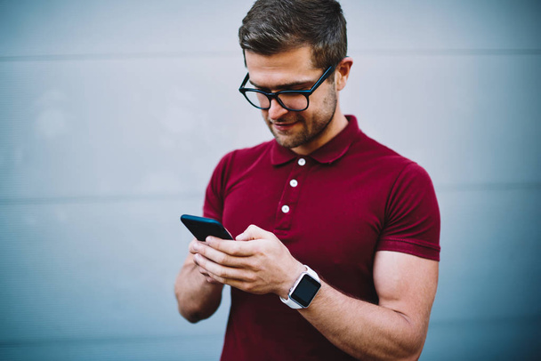 Young businessman in stylish eyeglasses standing along promotional background for your advertising and typing text message on smartphone.Handsome blogger publishing post in social networks on cellular - Foto, Imagen